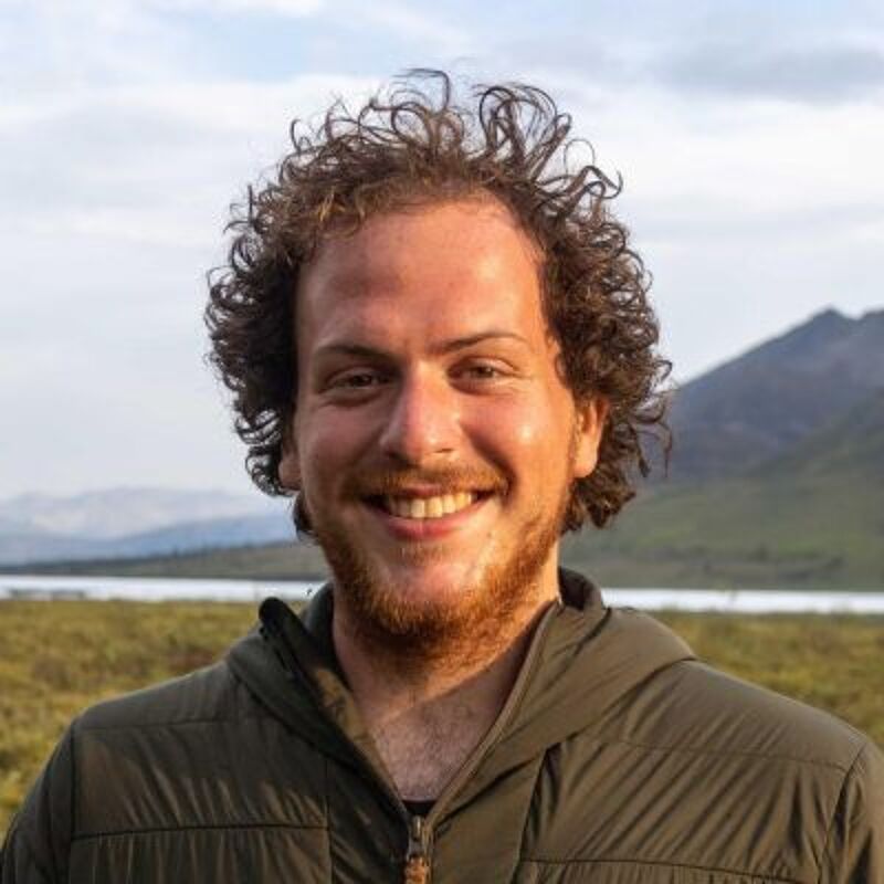 The image shows a man with curly brown hair and a beard, smiling at the camera. He is wearing a green jacket. The background features a lake, mountains, and a cloudy sky. The overall impression is of a person enjoying the outdoors.